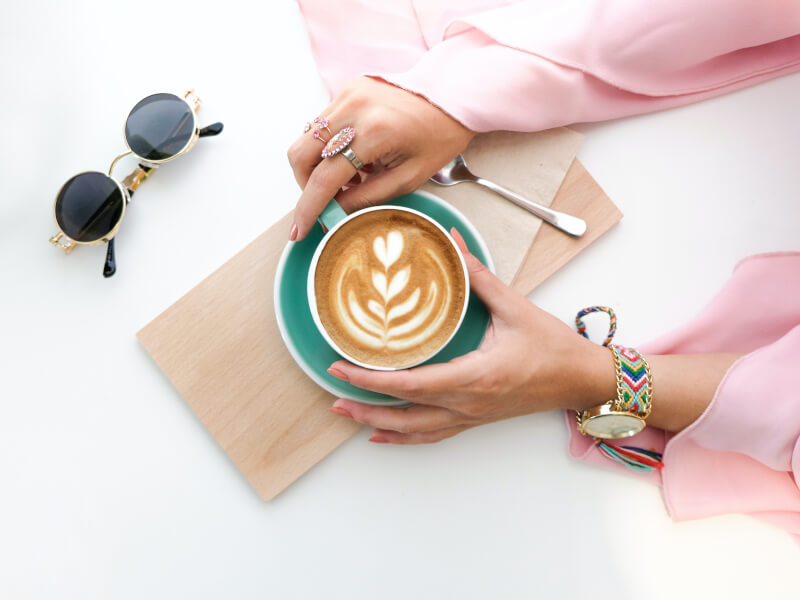 Shot from above of a person wearing a pink blouse and a beaded watch, holding a green coffee cup with latte art on the top